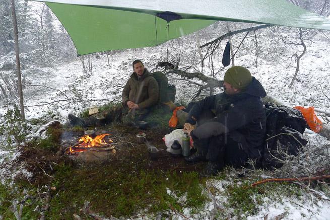 Two men sitting under a tarp with a fire surrounded by snow
