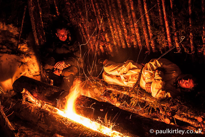 Two winter bushcraft guys in a classic lean-to shelter with textbook long-log fire
