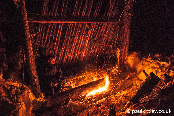 Lean to shelter illuminated by firelight of long log fire