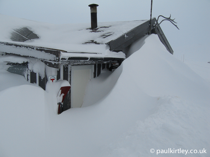 Hut with large snow drifts in front of it blocking the doorway