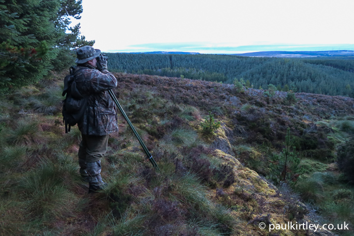 Man in drab clothing looking out over wild highland landscape using binoculars