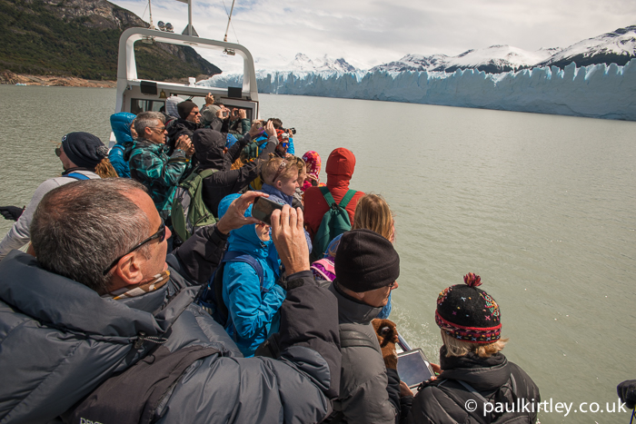 Many people on deck of a boat taking photographs of a glacier, causing the boat to lean towards the glacier