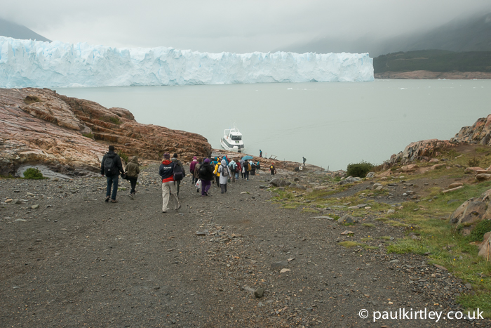 Tourists walking down a gravel track on a hill to the edge of a lake near a glacier