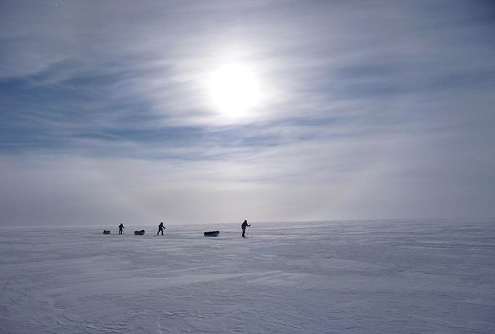 Three men hauling pulks in Antarctica, with milky sun through clouds