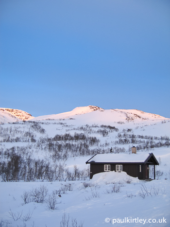 A small wooden hut agains the backrop of snow covered hills in Norway