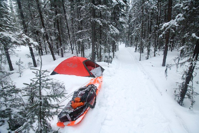 Red RAB tent and orange sled in white snow against black trees in the Yukon