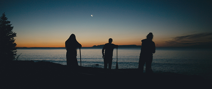 Three canoe people standing on a shoreline looking at sunset