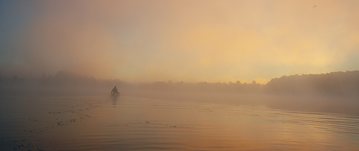 Man paddling a canoe on misty morning with orange light