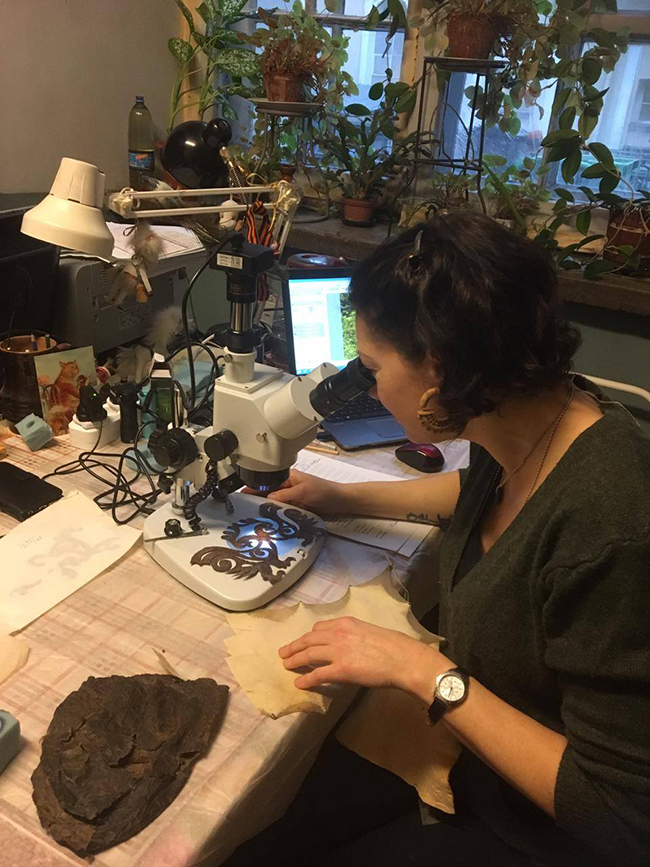 Woman looking through a microscope at an ornate sample of material, with desk lamp and laptop nearby
