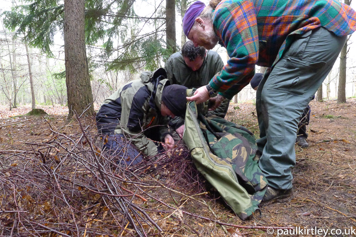 Man holding coat over pile of twigs while woman lights them with match