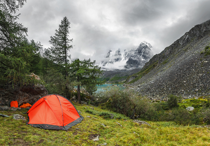 Orange tent in the mountains with grass, trees, rocks and snowy peaks in the distance.