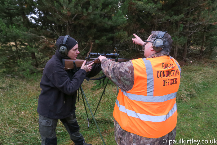 Man wearing bright orange tabard with "Range Conducting Officer" printed on back instructing woman with rifle.