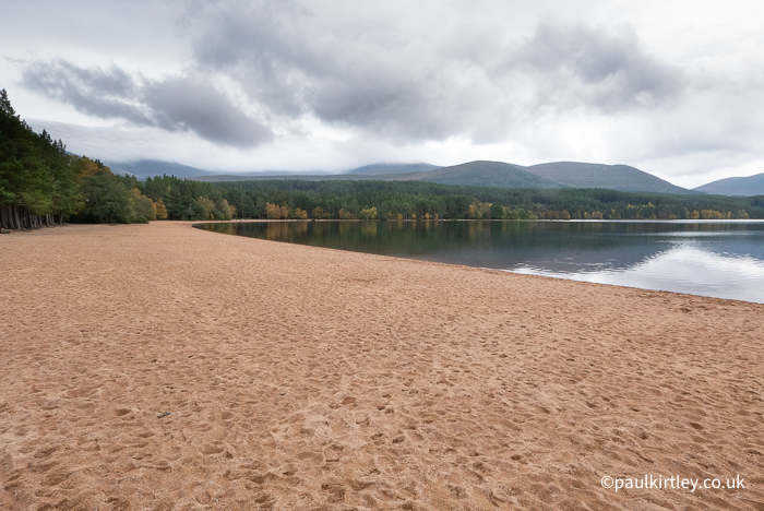 The large sandy beach of Loch Morlich, skirted by woodland, with the Cairngorms in the background