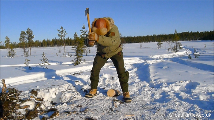 Man standing in open area on edge of boreal forest splitting firewood with an unusual technique