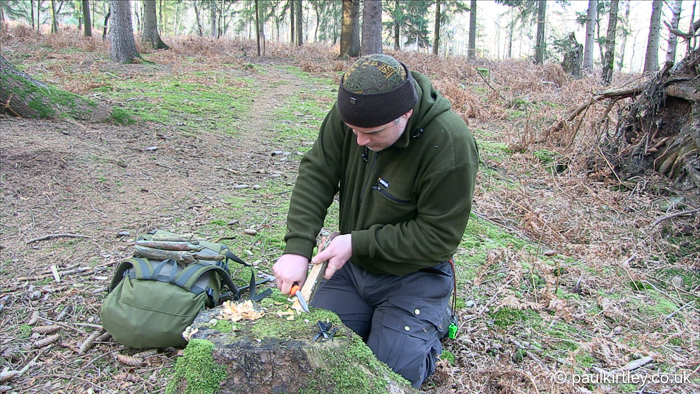 man shaving a block of wood with a knife in the forest