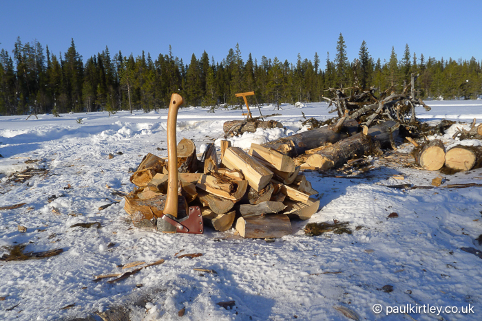 Snowy foreground with split logs and a small axe, against a background of coniferous boreal trees,