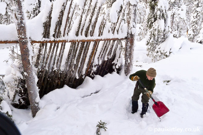 Man digging snow in front of a lean-to shelter.