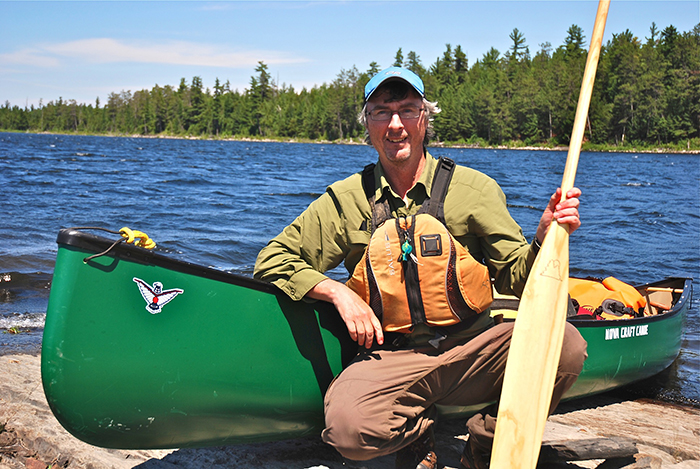 Man crouching in front of Nova Craft canoe, holding a canoe paddle