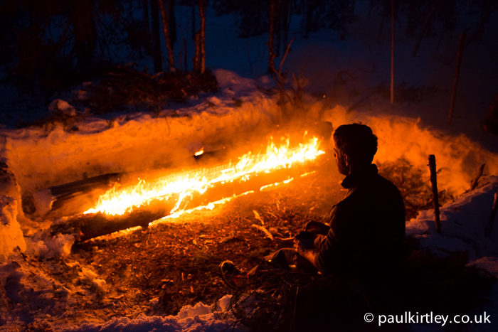 man sitting a little distance away from a long log fire