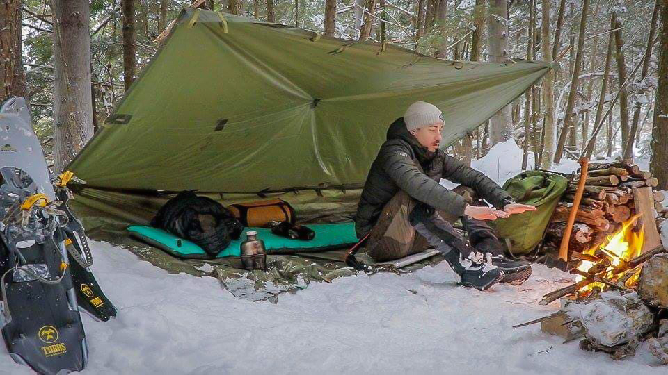 Man under tarp in snow with fire in front of him