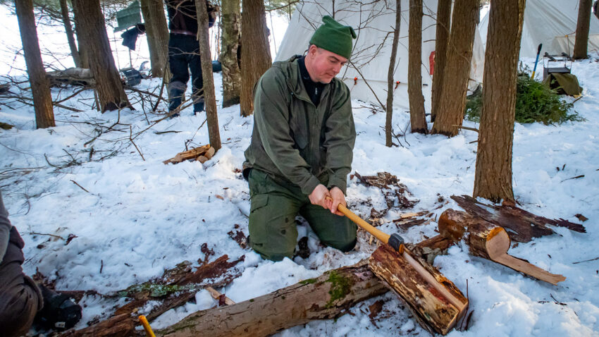 Man kneeling and splitting wood with an axe but no chopping block in a winter backcountry setting