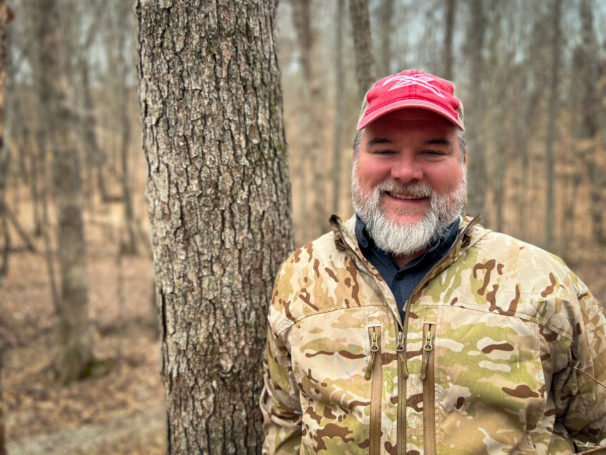 Happy , bearded American man in the woods, leaning against a tree, wearing camouflage pattern jacket and red cap.