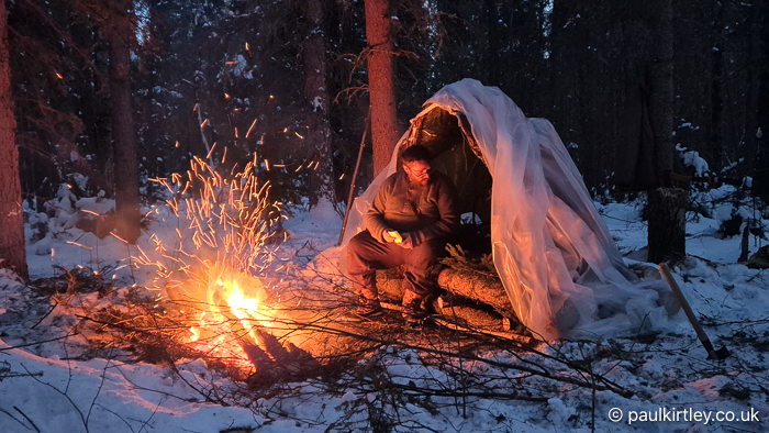 Man sitting in hybrid improvised shelter surrounded by dark forest but illuminated by fire in front of shelter