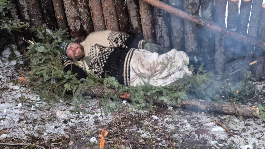 Warmly clothed man lying on spruce bough bed in an improvised northern forest shelter